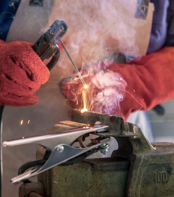 A man welder with safety helmet working with arc welding machine in the workshop, sideways flashes. Male hands in gloves fix a piece of iron with sparks fly in different directions.